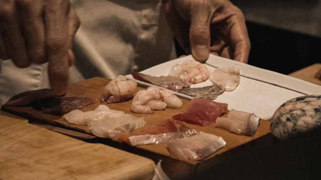 Close-up shot of a chef presenting a wooden platter displaying an array of fresh raw sashimi slices, including tuna, white fish, and sweet prawns.
