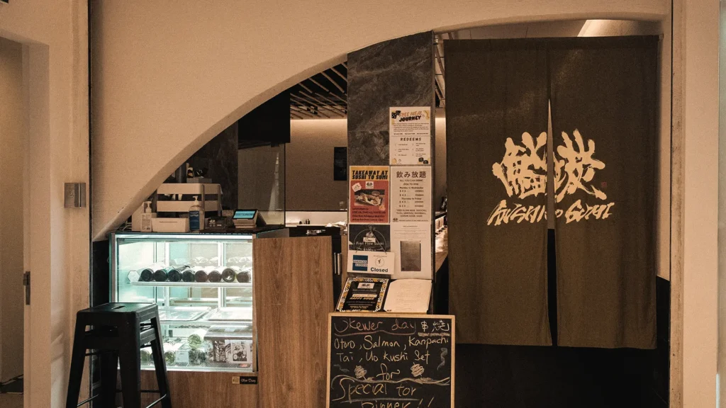 Eye-level shot of a Japanese restaurant entrance decorated with a traditional dark noren curtain, an illuminated wine cooler, and a handwritten chalkboard menu easel.