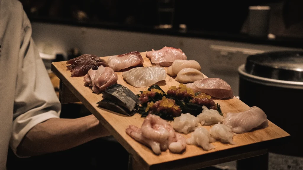 Medium close-up shot of a chef holding a wooden tray filled with a freshly prepared selection of raw seafood cuts, including scallops, sweet shrimp, and sliced fish.