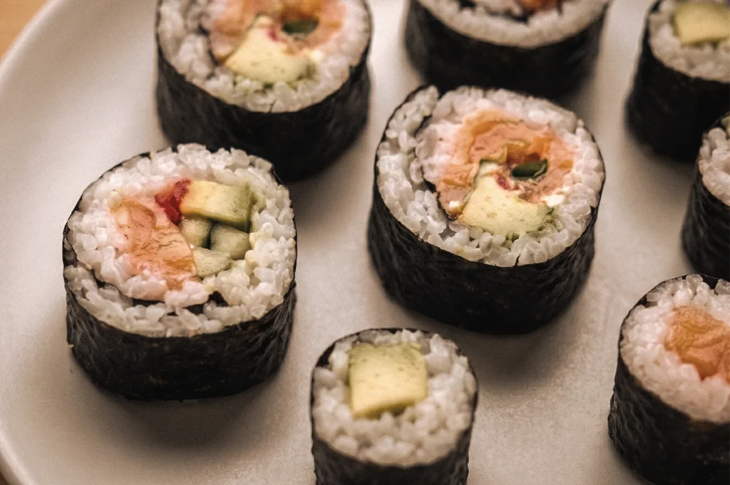 Close-up 45-degree angle of traditional Japanese hosomaki and futomaki sushi rolls on a white plate, featuring nori-wrapped rice with salmon, cucumber, and avocado fillings, highlighting the difference between thin and thick sushi rolls.