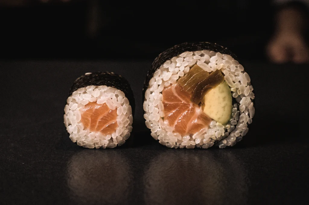 Eye-level macro view of hosomaki and futomaki sushi roll cross-sections showing salmon, avocado, cucumber, and seaweed fillings, illustrating the size and filling differences between thin and thick Japanese sushi rolls.