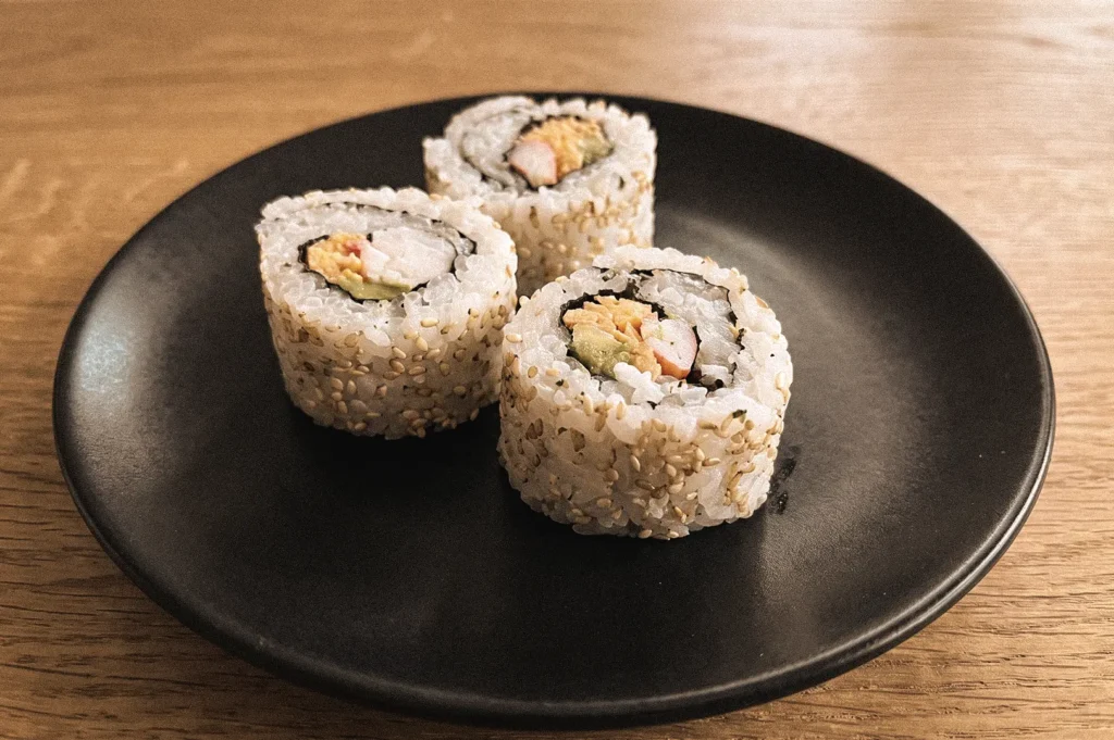 Three sesame-coated uramaki inside-out sushi rolls on a black plate, showing rice on the outside with crab, avocado and cucumber filling wrapped in hidden nori, photographed from a slight high-angle close-up view.