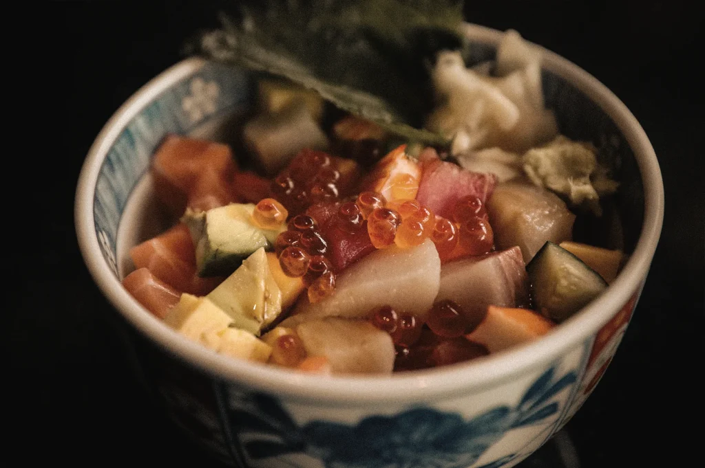 Bara chirashi sushi bowl featuring diced salmon, tuna, scallops, avocado, cucumber, and ikura salmon roe over vinegared sushi rice, photographed from an eye-level macro angle to highlight the colorful scattered sushi presentation.