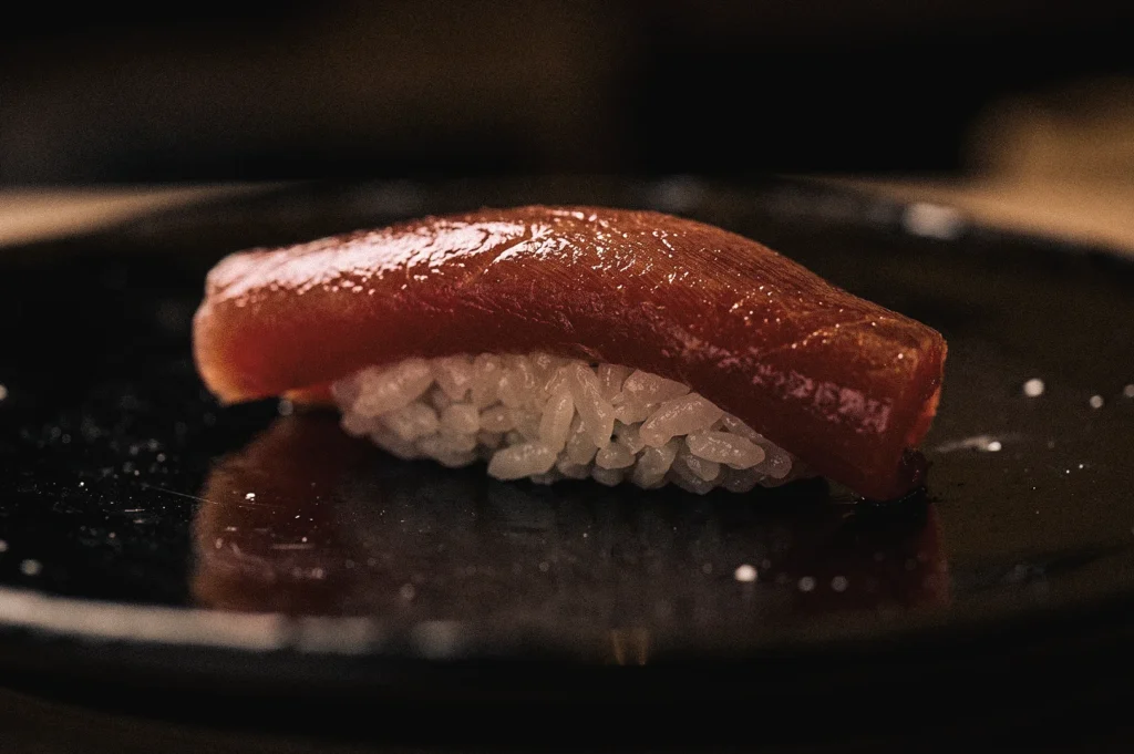Close‑up shot of glossy akami tuna nigiri marinated in soy sauce (zuke), resting on seasoned rice, capturing Edomae sushi’s Tokyo tradition of hand‑formed nigiri and flavour‑enhancing preparation.