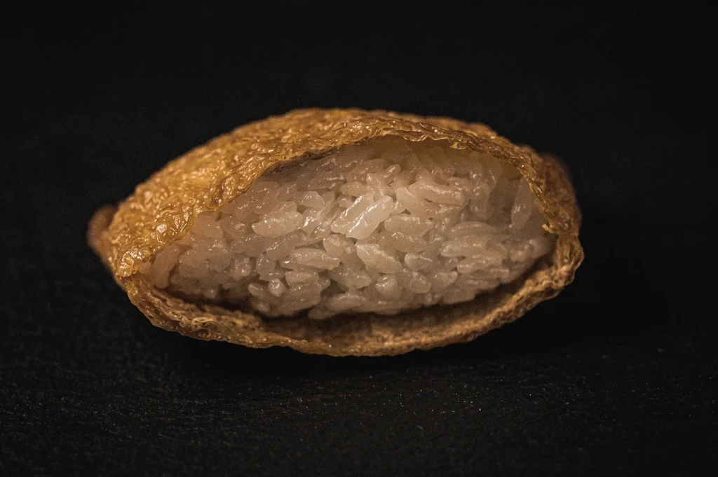 Low-angle close-up of a single piece of inari sushi showing sushi rice stuffed inside a sweet seasoned fried tofu pouch against a dark background.
