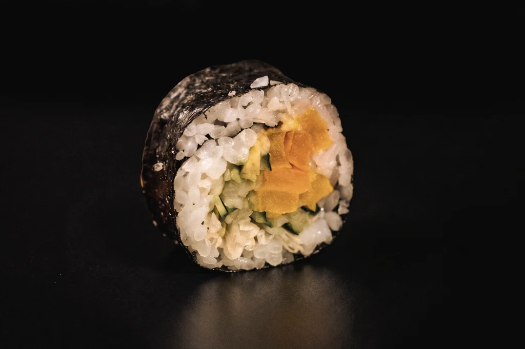 Front-facing macro photograph of a single vegetable maki sushi roll, highlighting nori seaweed, seasoned sushi rice, and fresh fillings, illustrating the structure of traditional Japanese rolled sushi.