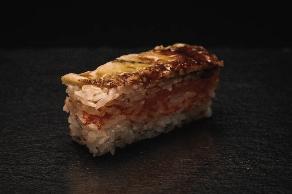 Side-angle close-up of aburi oshizushi topped with flame-seared seafood and sauce, highlighting modern Japanese pressed sushi with caramelized flavors and textured vinegared rice.