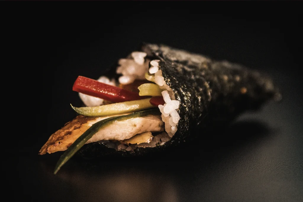 Close-up macro view of a temaki sushi hand roll filled with rice, grilled protein, cucumber, and red pepper, photographed from a low angle to highlight the crisp nori cone and fresh Japanese sushi ingredients.