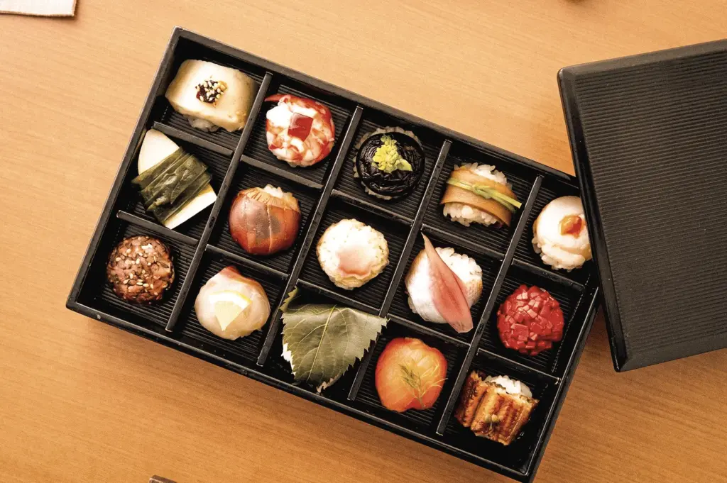 Top-down flat lay shot of an assorted Temari Sushi box featuring premium seafood, tamago, vegetables, and seasonal toppings arranged in a traditional Japanese bento-style presentation.