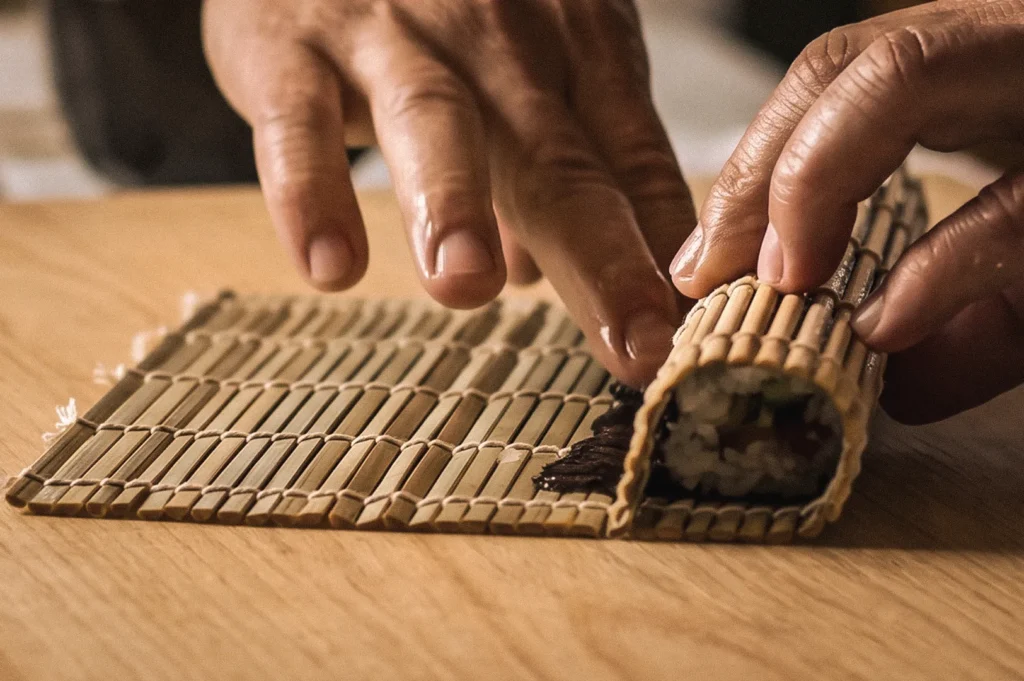 Close-up side-angle photograph of sushi being rolled with a traditional bamboo makisu mat, demonstrating the preparation of authentic Japanese hosomaki and futomaki sushi rolls.