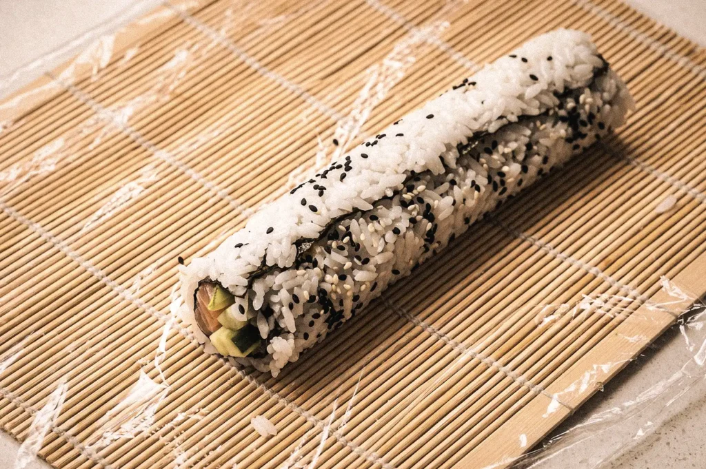 Fresh uramaki inside-out sushi roll on a bamboo rolling mat during preparation, featuring sushi rice on the outside, nori hidden inside, avocado filling, and black sesame seeds, photographed from a high-angle view.
