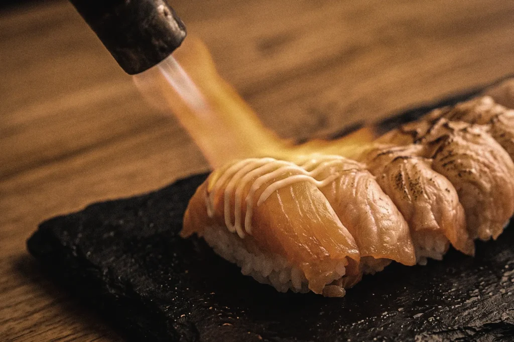 Chef torch-searing aburi salmon nigiri sushi with a handheld flame, captured in a close-up diagonal overhead angle, highlighting the caramelization process and smoky flavor development of authentic Japanese aburi sushi.