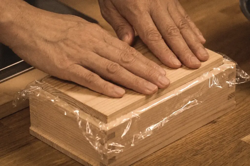Close-up overhead shot of a wooden oshizushi mold being pressed by hand during the preparation of Battera sushi, Osaka's traditional pressed mackerel sushi.