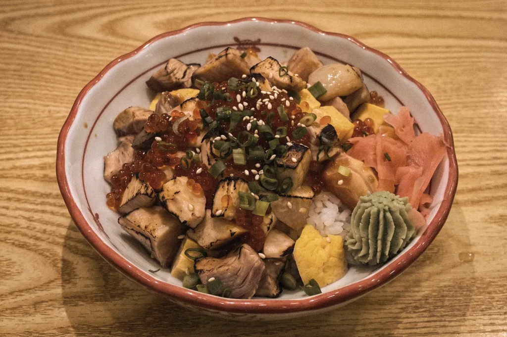 Edomae-style chirashizushi topped with diced sashimi, ikura salmon roe, tamago, scallions, sesame seeds, pickled ginger, and wasabi over seasoned sushi rice, photographed from a 45-degree overhead angle highlighting traditional Japanese scattered sushi.
