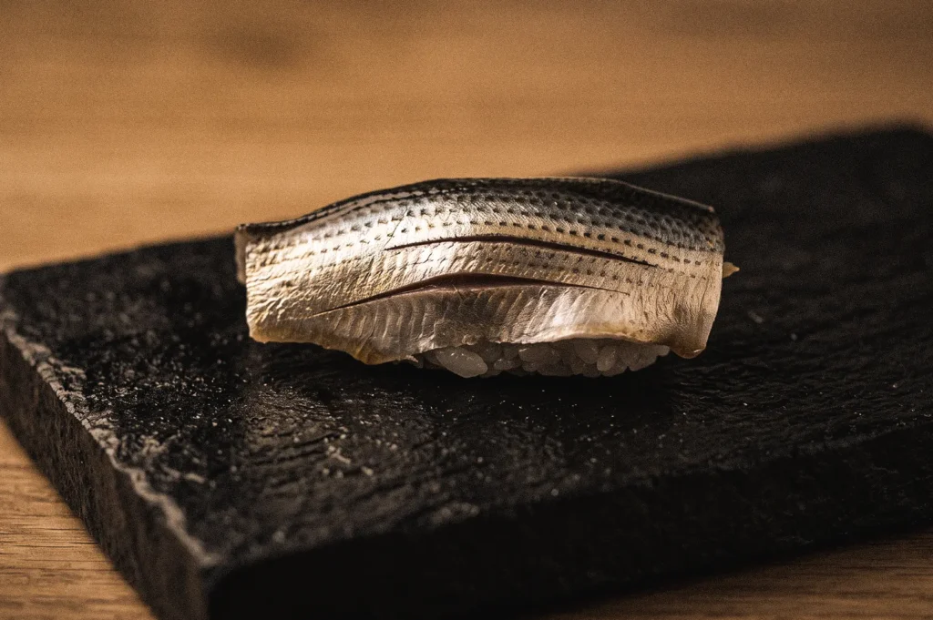 Close-up low-angle macro shot of traditional Edomae kohada nigiri sushi, featuring vinegar-cured gizzard shad over seasoned rice, showcasing classic Edo-style sushi preparation techniques.
