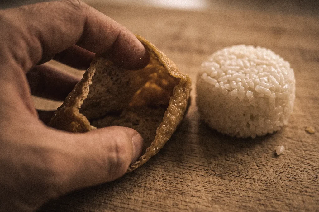 Close-up macro shot of a hand opening a seasoned fried tofu pouch (aburaage) beside vinegared sushi rice while preparing traditional Japanese inari sushi.