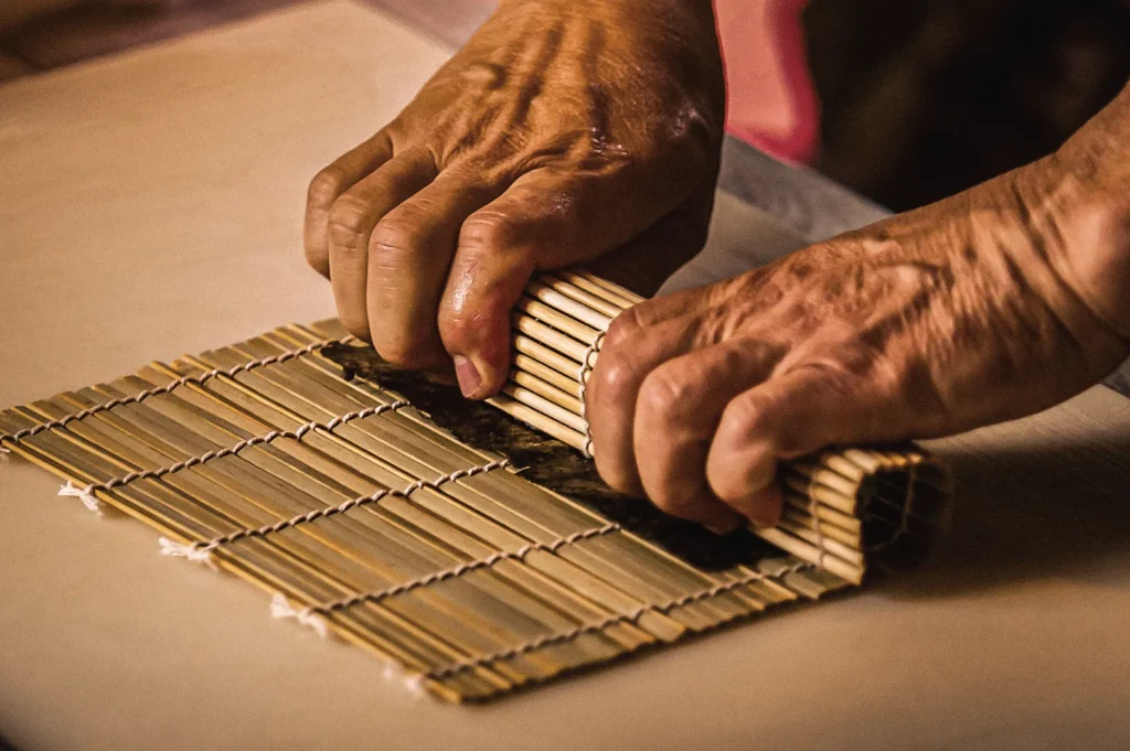 High-angle close-up of hands using a bamboo makisu mat to roll maki sushi, demonstrating the traditional Japanese sushi-making technique used to create nori-wrapped rolled sushi.