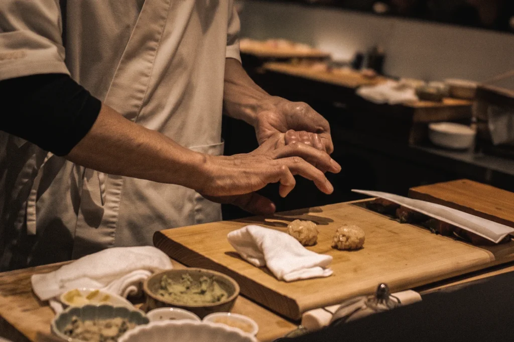 Over-the-counter medium close-up of a sushi chef hand-shaping nigiri sushi on a wooden board, demonstrating traditional Japanese sushi-making techniques and authentic omakase craftsmanship.
