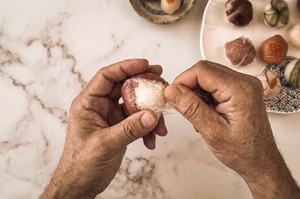 Overhead close-up shot of hands using plastic wrap to shape traditional Temari Sushi, a Japanese ball-shaped sushi made with vinegared rice and sashimi topping on a marble countertop.
