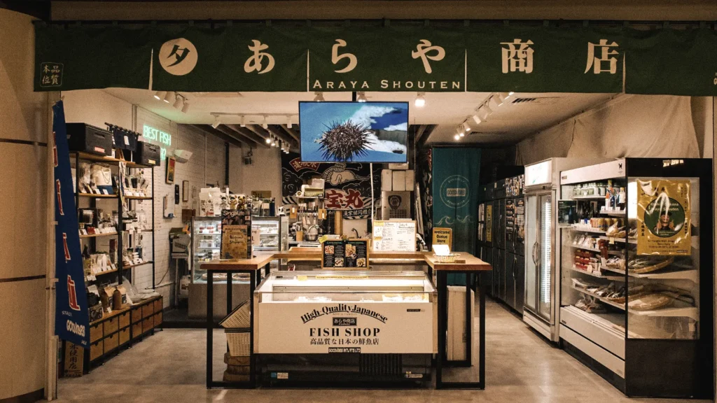 Front view of Araya Shouten, a Japanese fish shop with refrigerated seafood displays, shelves of specialty products, and a large sign bearing the shop name in Japanese and English above the entrance.