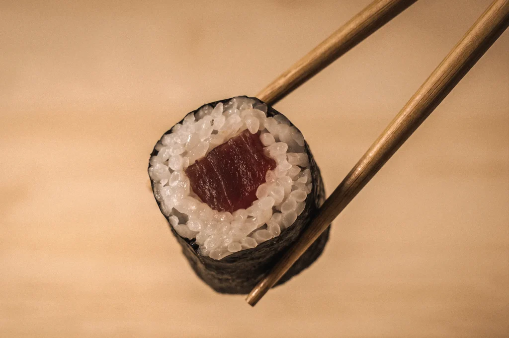 Macro close-up of a traditional tekkamaki hosomaki sushi roll held with chopsticks, featuring a single filling of lean tuna wrapped in sushi rice and nori, demonstrating the classic thin-roll style in Japanese sushi culture.