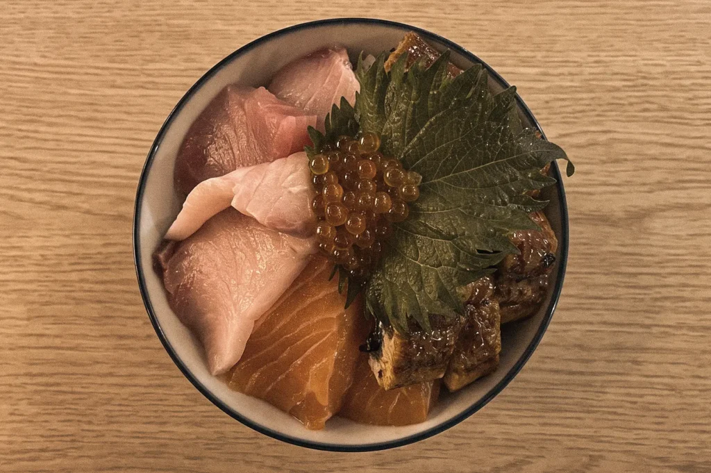 Edomae-style chirashizushi viewed from a top-down angle, featuring salmon sashimi, tuna, white fish, ikura salmon roe, grilled eel, and shiso leaf arranged over vinegared sushi rice in a traditional Japanese scattered sushi bowl.