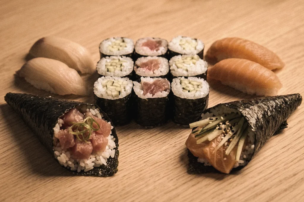 Fresh Japanese sushi assortment featuring temaki hand rolls, maki sushi rolls, and nigiri sushi arranged on a wooden table, photographed from a 45-degree angle to illustrate the differences between temaki, maki, and nigiri sushi.