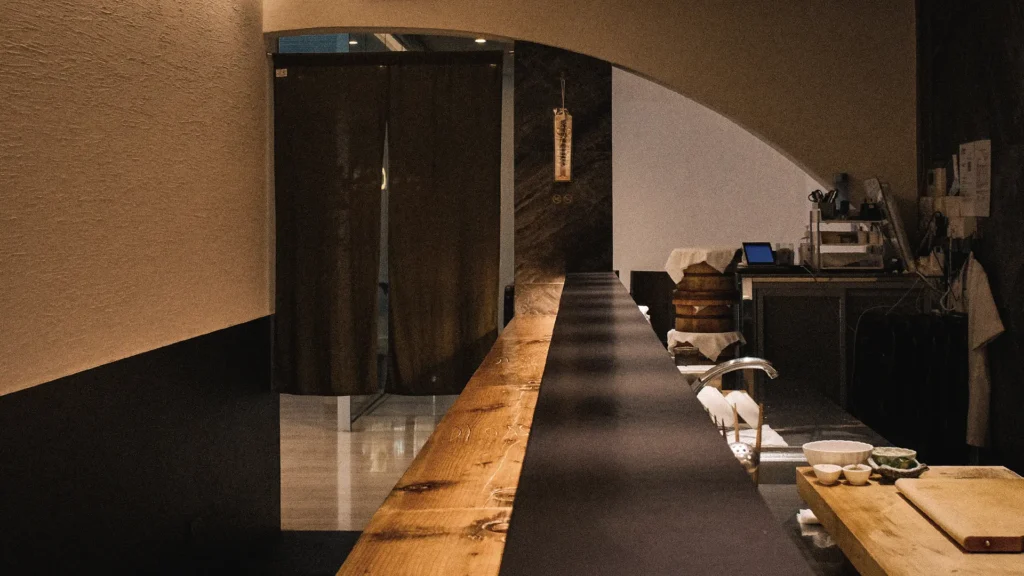 Interior view of the standing counter at Sushi to Sumi, featuring a long wooden counter running the length of the narrow dining space. The minimalist restaurant is softly lit with warm tones, with the open preparation area visible behind the counter and dark curtains hanging at the entrance to the kitchen area.