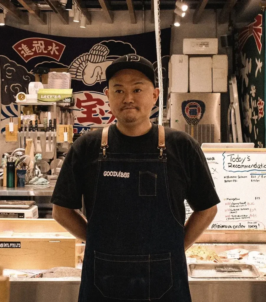 Araya-san stands behind a fresh seafood display at Araya Shouten, a Japanese fish shop. Refrigerated cases filled with fish and seafood line the foreground, while recommendation boards, refrigerated storage units, and fishing-themed décor are visible throughout the shop interior.