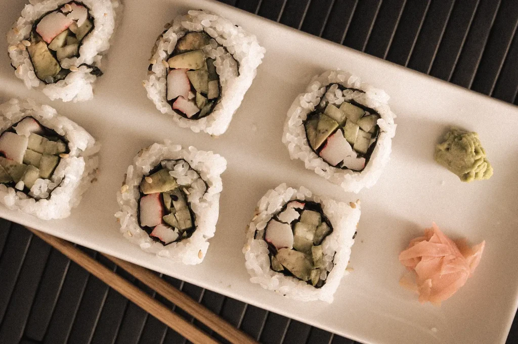 Overhead view of sliced California roll inside-out sushi rolls with cucumber, imitation crab and hidden nori, served on a white rectangular plate with wasabi and pickled ginger.