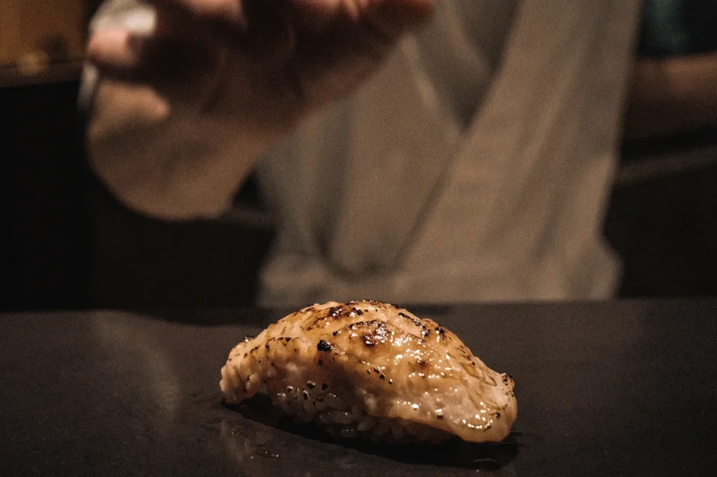 Premium aburi nigiri sushi with a flame-seared topping resting on seasoned sushi rice, photographed from an eye-level close-up angle at a Japanese sushi counter, emphasizing glossy texture, charred details, and artisanal preparation.