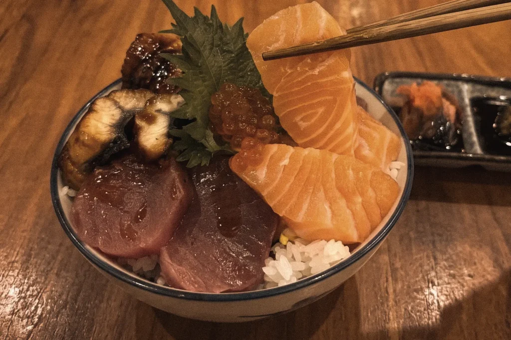 Chopsticks lifting a slice of salmon sashimi from a chirashizushi bowl topped with tuna, ikura salmon roe, grilled eel, and shiso leaf over vinegared sushi rice, photographed from a 45-degree close-up angle to demonstrate how chirashi sushi is enjoyed.