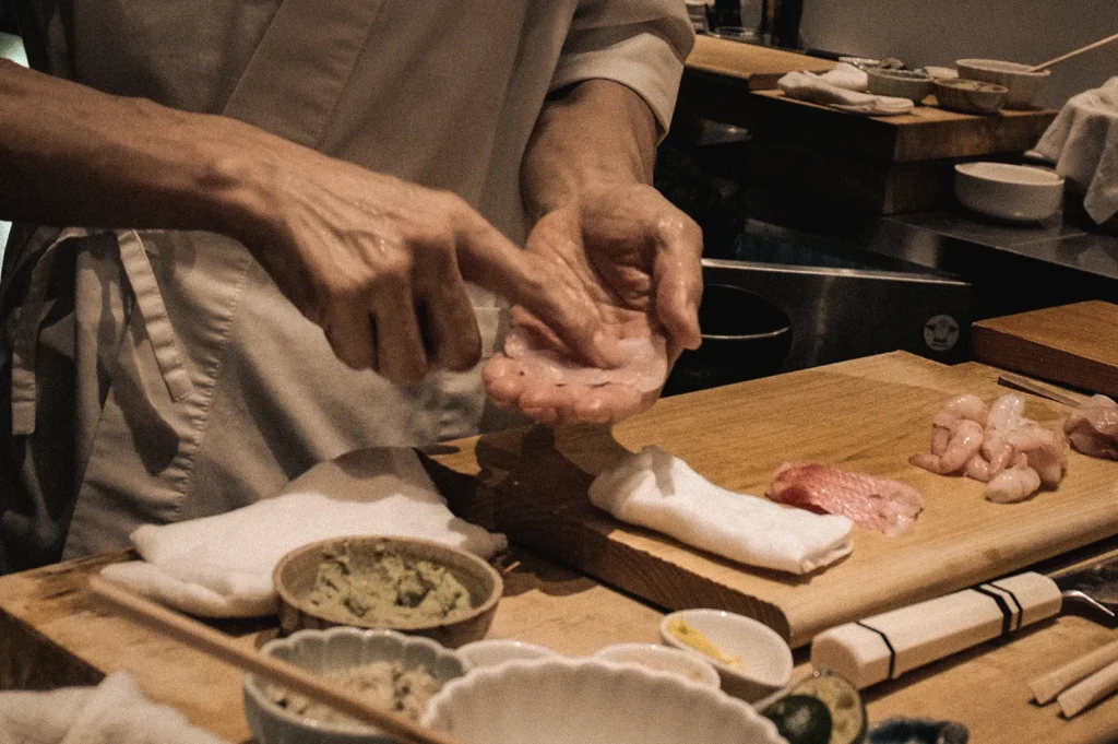 Close-up action shot of a sushi chef preparing Edomae nigiri at a sushi counter, demonstrating traditional fish handling and sushi-making techniques rooted in Edo-period sushi culture.