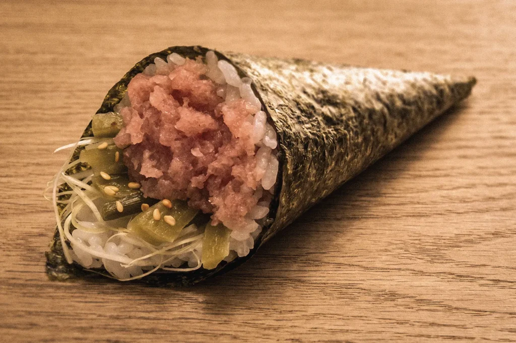 Close-up side-angle view of a negitoro temaki hand roll filled with fatty minced tuna, sushi rice, nori, scallions, and sesame seeds, highlighting a classic Japanese temaki sushi filling served fresh for immediate eating.