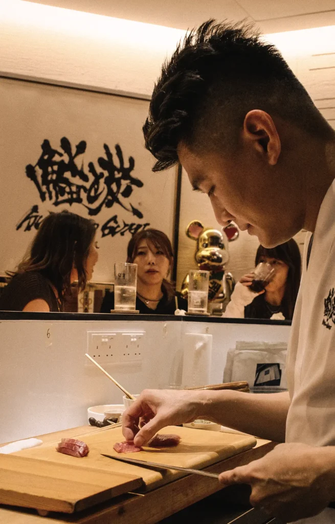 Side-angle close-up of a chef preparing premium tuna sushi at Sushi to Sumi, an intimate omakase restaurant, with guests dining at the counter in a warm Japanese-inspired setting.