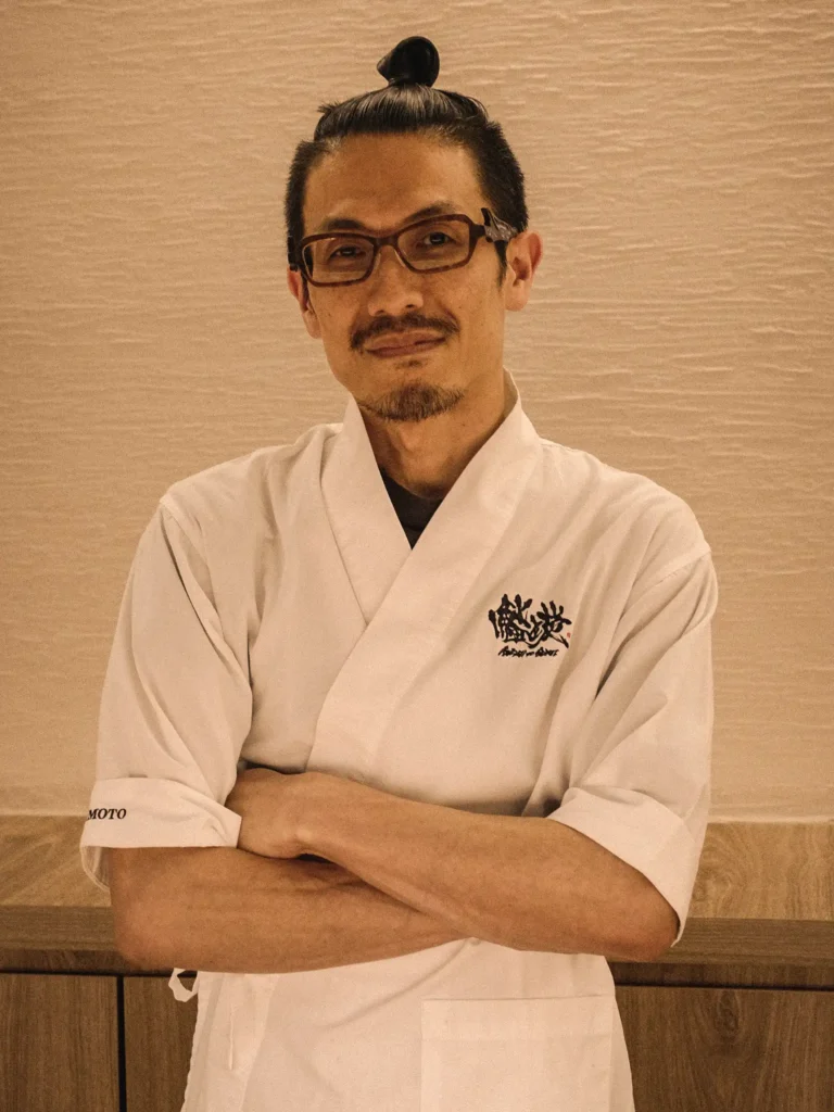 Hashimoto Yasuo, who runs the counter at Sushi to Sumi, stands with arms folded wearing a white chef coat and glasses against a light textured wall.